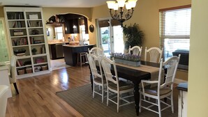 A dining area with a wooden table and white chairs, a bookshelf, and a kitchen in the background. - Charming Floridian Home with pool located close to Lauderdale by the Sea. (Fort Lauderdale)