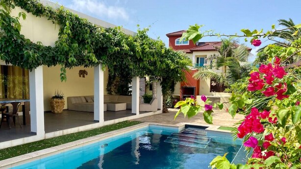 A swimming pool with a covered patio area, surrounded by lush greenery and vibrant flowers.