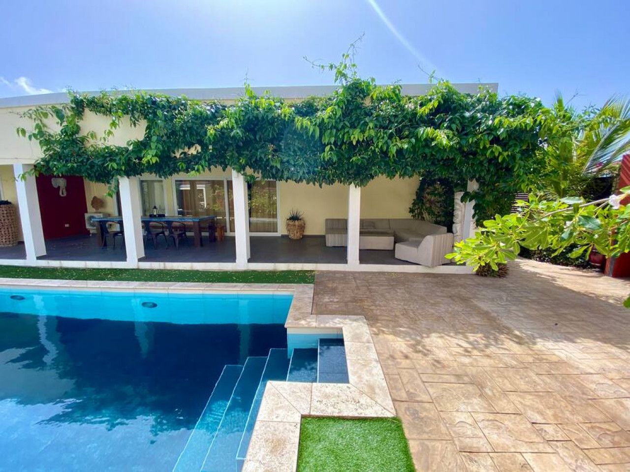 Une piscine avec une terrasse carrelée, un patio couvert avec des sièges et une pergola ornée de plantes grimpantes.