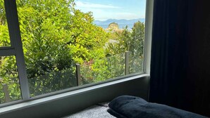 Un lit recouvert d’une couvre-lit à motifs floraux, une fenêtre donnant sur un paysage verdoyant et montagneux, et un rideau foncé partiellement tiré.