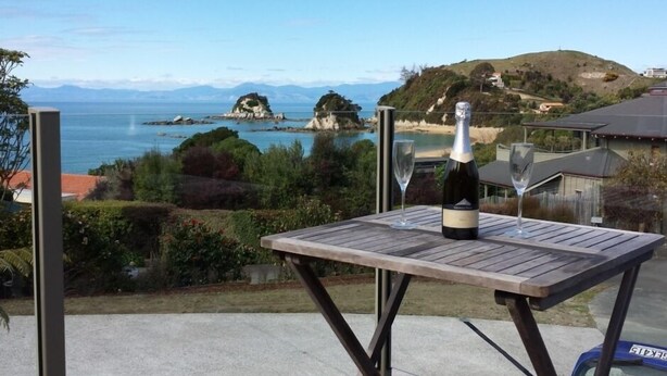 Une table avec une bouteille de vin et un verre, placée sur un balcon donnant sur un paysage côtier avec des collines et une vue sur la mer.