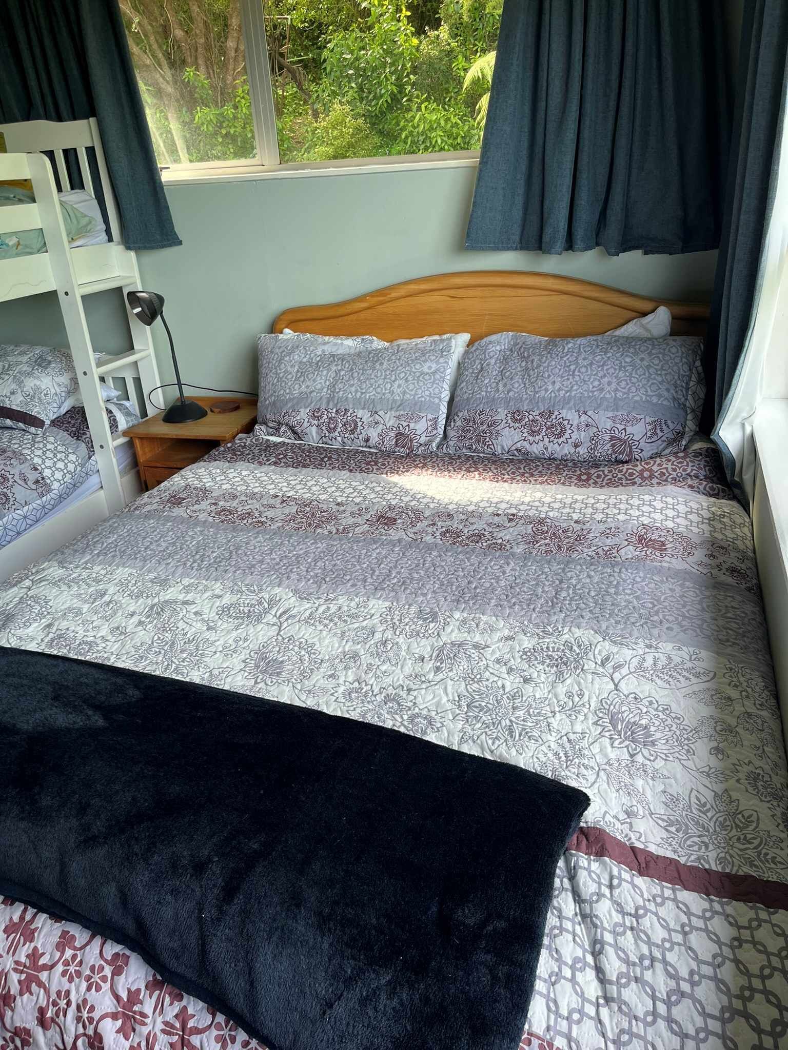 Ein Schlafzimmer mit einem hölzernen Bett, gemusterten Betttextilien, einem Nachttisch, einem Schreibtisch und einem Fenster mit Vorhängen.