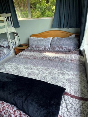 A bedroom with a wooden bed, patterned bedding, a nightstand, a desk, and a window with curtains. - Little Kaiteriteri Rose's bach -3min walk to beach (Kaiteriteri)