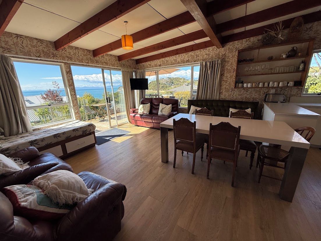 A spacious living area with a large window offering a view of the sea, a dining table with chairs, and a sofa with cushions.