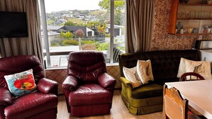 Un salon avec un fauteuil en cuir, un canapé vert et une table à manger en bois. Une grande fenêtre offre une vue sur un quartier résidentiel.