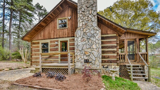 Une cabane en rondins avec une cheminée en pierre, une terrasse en bois et un sentier de pierre menant à l’entrée.