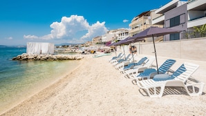A beach with white lounge chairs and umbrellas, a rocky breakwater, and buildings in the background. - Villa Four Brothers - Exclusive Apartment (Podstrana)