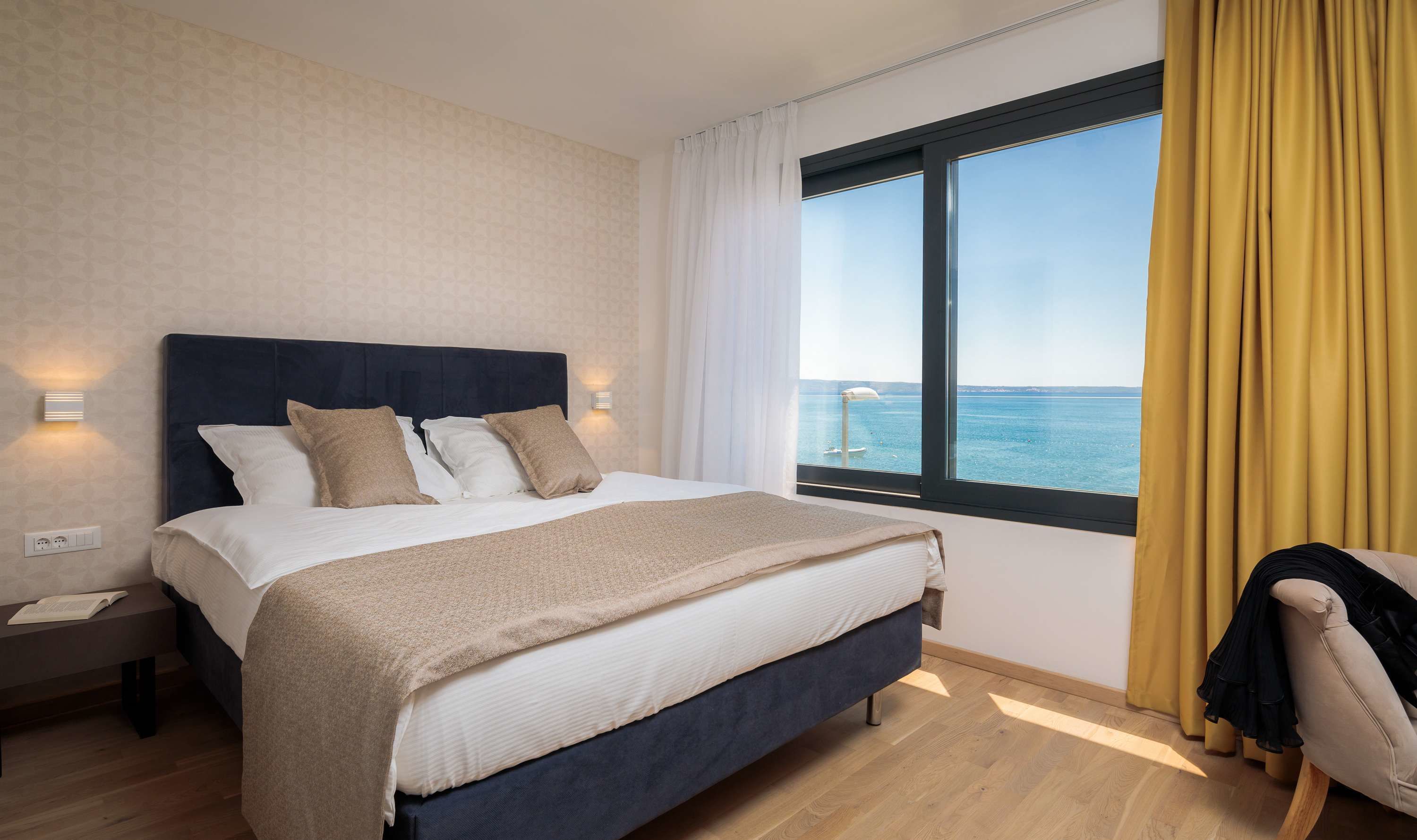 Een netjes opgemaakt bed met een donker hoofdbord, een nachtkastje met een lamp, en uitzicht op de zee door een groot raam met gele gordijnen.