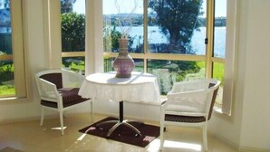 Quarto com uma mesa redonda e duas cadeiras brancas, janelas amplas com vista para um lago e árvores, e um vaso sobre a mesa.