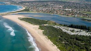 Vista aérea de uma área costeira com uma praia, um rio e uma cidade densamente povoada ao fundo.