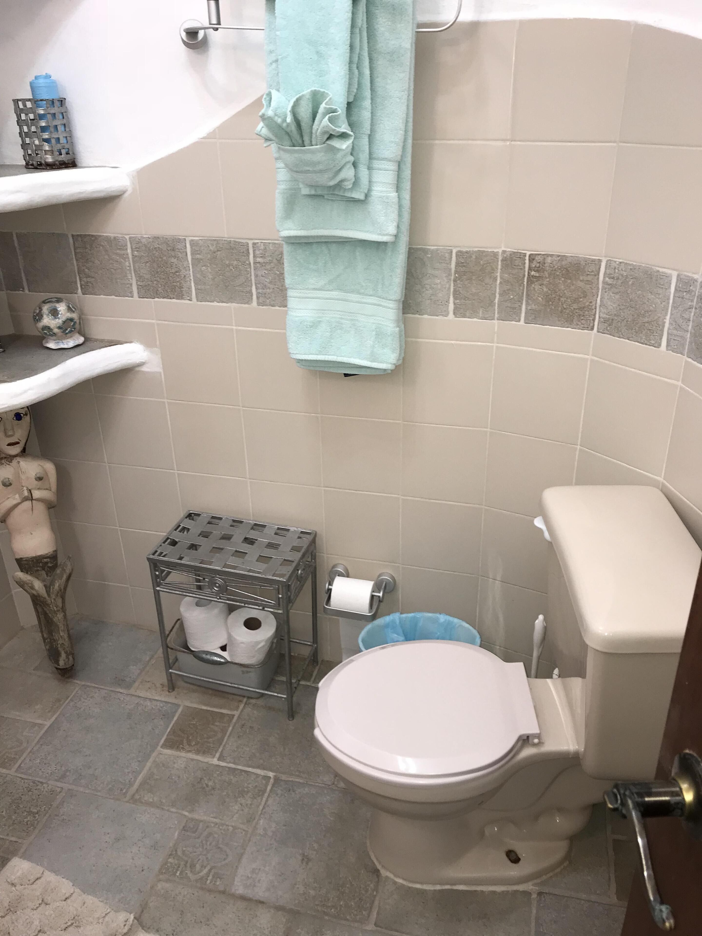 A bathroom with a toilet, a sink, a towel rack, a shelf with toiletries, and a trash bin.