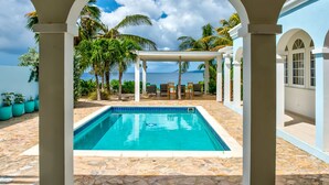 A swimming pool with a stone-tiled deck, surrounded by white pillars and a covered seating area with chairs and a table.