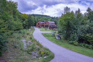 Une grande maison à deux étages, dotée d’une allée sinueuse, entourée d’une forêt dense et de collines.