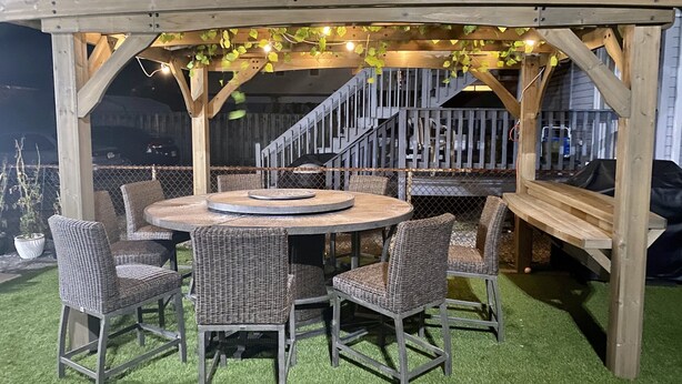A wooden pergola with string lights and ivy, a round table with chairs, and a fenced outdoor area.
