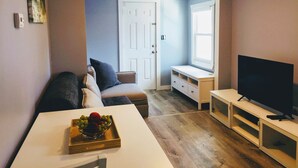 A compact living space with a white sofa, a coffee table with a fruit bowl, a TV on a stand, and a door leading to another room.