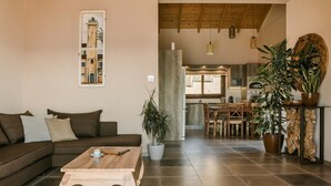 Una sala moderna con un sofá marrón, una mesa de centro de madera y una zona de comedor con una mesa y sillas de madera. Hay plantas en macetas y un espejo grande en el lado derecho.