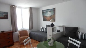 Een kleine kamer met een bed, een stoel, een tafeltje en een bureau. Er hangt een schilderij aan de muur en er is een raam met gordijnen.