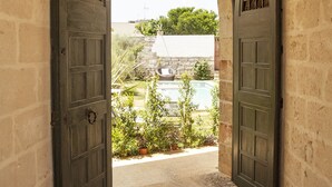 An arched doorway with two dark wooden doors leading to a courtyard with a swimming pool and greenery. - new apt with private garden, all comforts, WI-Fi & AC, in Salento's hearth (Diso)
