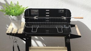 En grill med træstel og metalrist, placeret på en træterrasse foran en hvid væg.