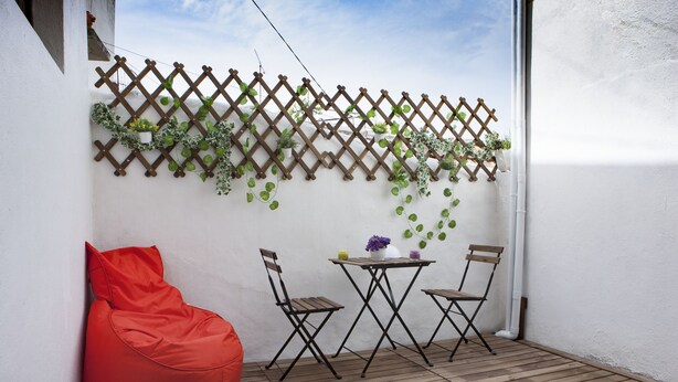En balkon med trægulv, en rød oppustelig pude, to sammenklappelige stole, et lille bord med en vase blomster og en espalier væg med slyngplanter.