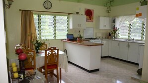 A kitchen with a dining table, chairs, a clock, and a window with blinds. - Malau Lodge (Nuku'alofa)