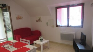 A small, well-lit room with a red sofa, a white table, a small white table, a radiator, and a television on a black stand. - STUDIO RENTAL DOMAINE DE LA BAUME (CENDRAS)