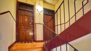 Una escalera de madera con barandilla roja y otra de metal oscuro, que conduce a una puerta de madera con un aldabón de latón.