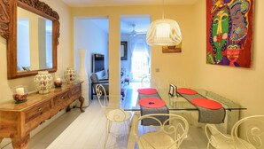 Un comedor con una mesa de vidrio, sillas blancas y un cojín rojo. Se observa un aparador de madera con adornos y un espejo grande. En la pared hay un cuadro de colores, y se ve un ventilador de techo.