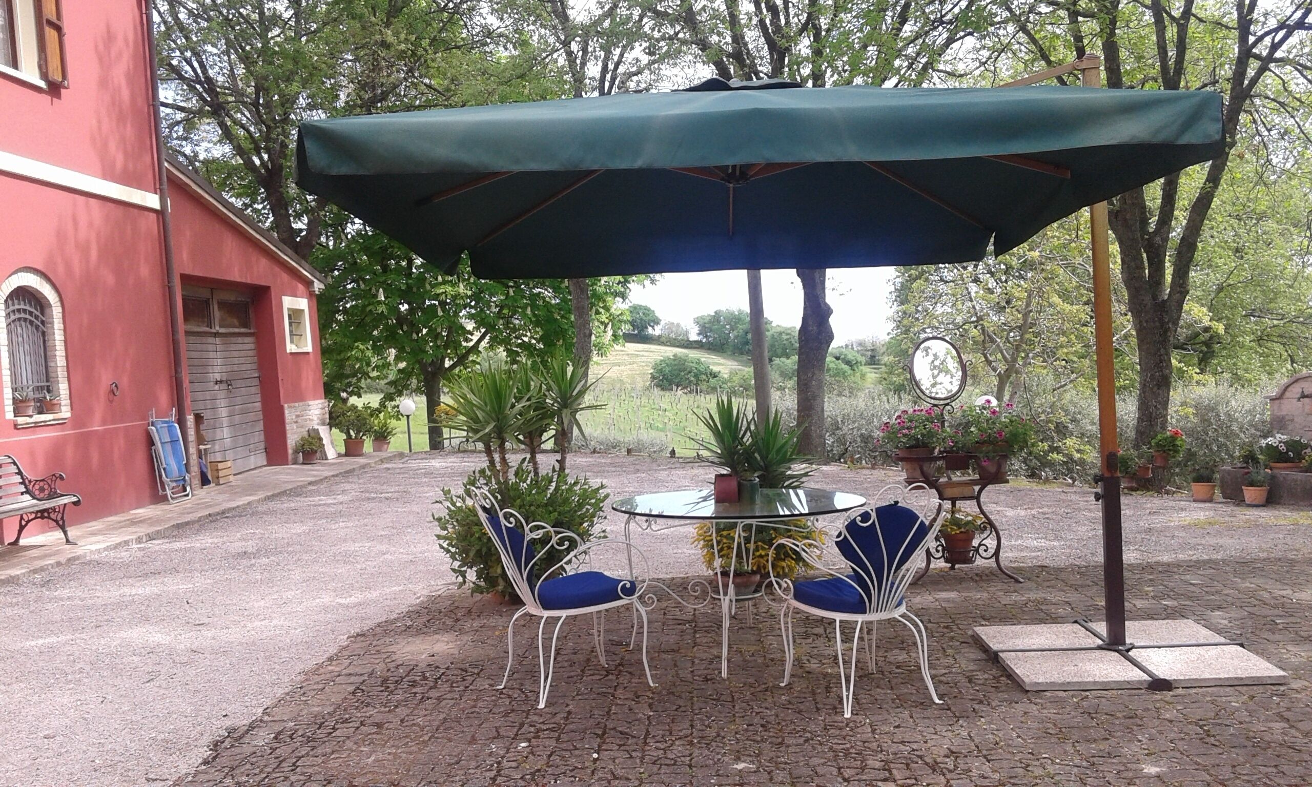An outdoor patio with a table and two chairs under a green umbrella, surrounded by potted plants and a red building.