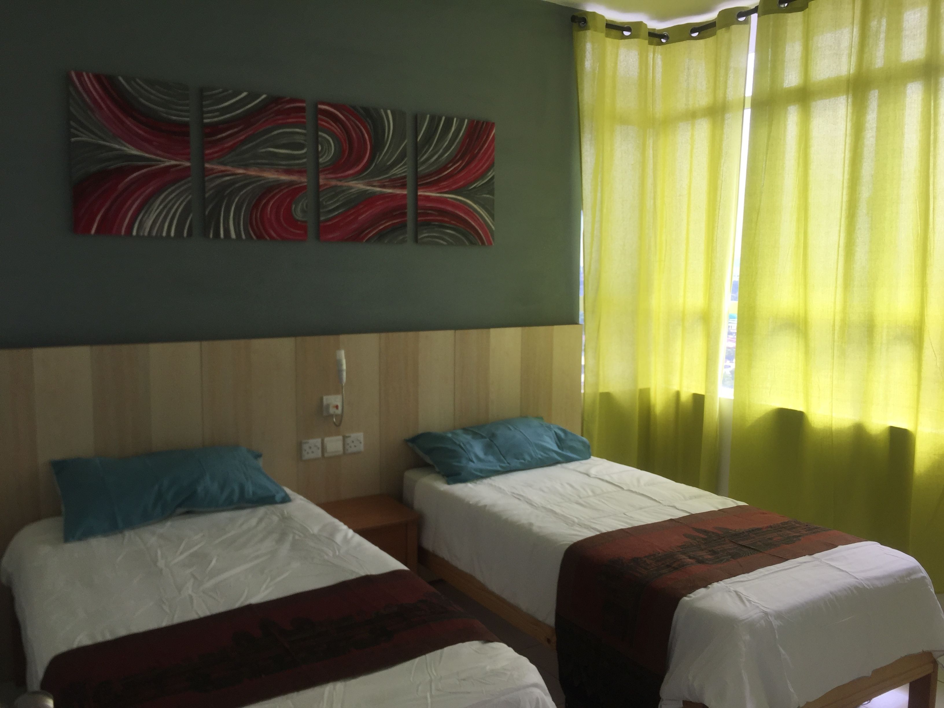 A hotel room with two single beds, a wall-mounted power outlet, and a window with yellow curtains.