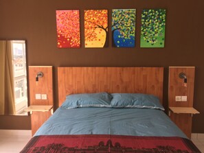 Una habitación de hotel con cabecera de madera, cama con ropa de cama azul y cuatro cuadros coloridos en la pared.