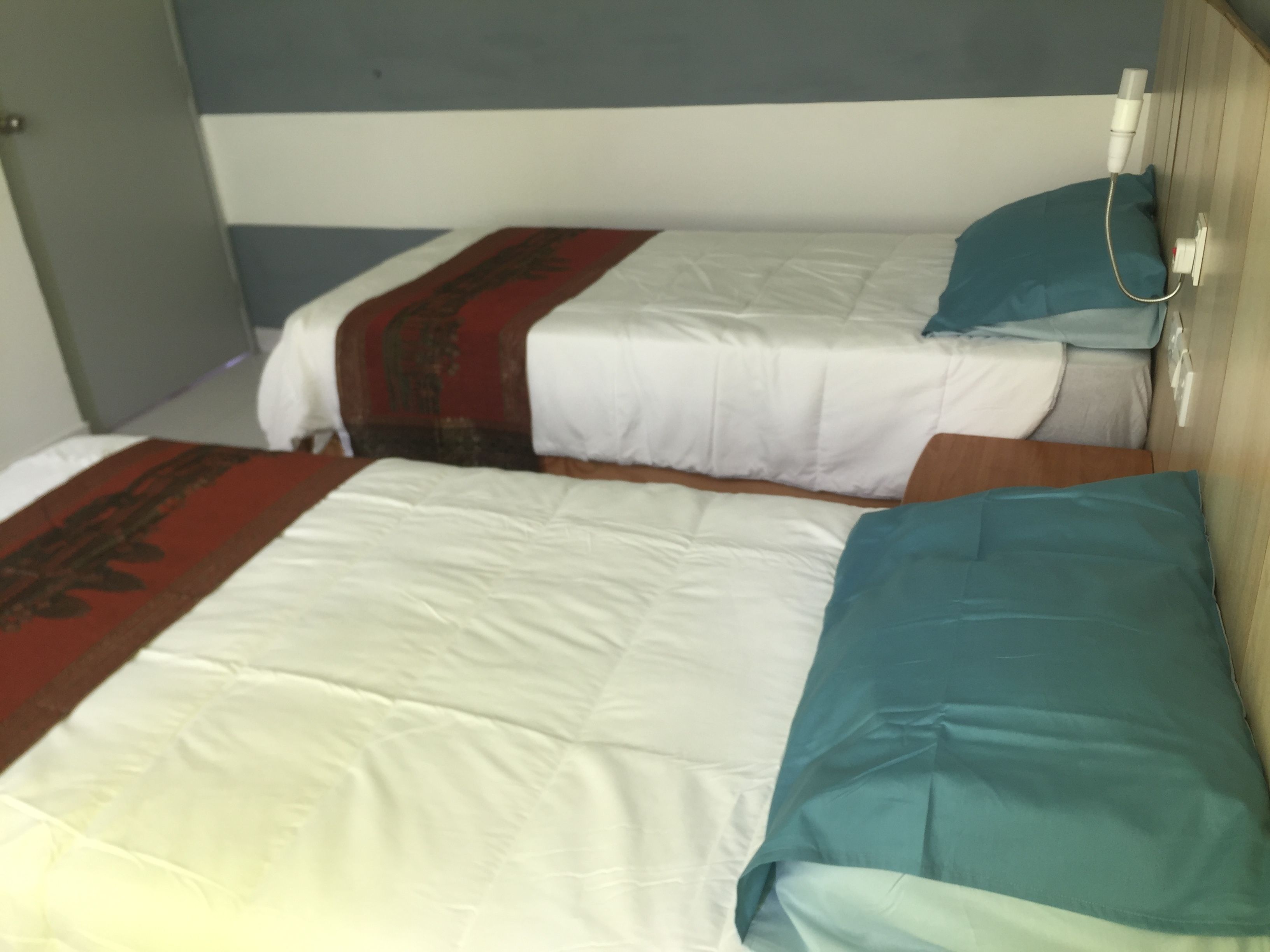 Two beds with white and teal bedding, a wooden headboard, and a bedside table with a lamp.