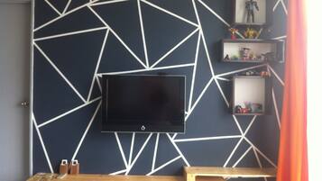Een kamer met een aan de muur gemonteerde televisie, een houten bank en planken met diverse spullen.