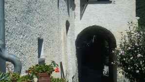 Ein Steinweg führt zu einem Gebäude mit rustikaler Fassade, einem Bogenportal und Kübelpflanzen am Wegesrand.