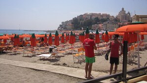Ein Strand mit Reihen von orangefarbenen Sonnenschirmen und Liegestühlen, ein Mann in einem roten Hemd und ein Gebäude im Hintergrund.