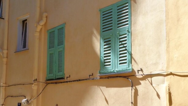Un bâtiment doté de nombreuses fenêtres, dont certaines sont équipées de volets, et d’une porte surmontée d’un portail en métal.