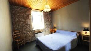 A bedroom with a stone wall, a wooden ceiling, a bed with a blue bedspread, a chair, a nightstand with a lamp, and a window letting in natural light. - Winetourism in the heart of Bordeaux (Margueron)