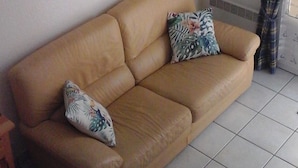 Una sala de estar con un sofá beige, dos cojines estampados, un cuadro floral en la pared y una ventana con cortinas a cuadros.