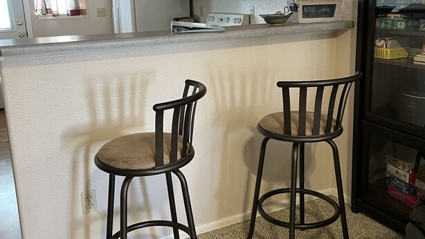 A kitchen with wooden cabinets, a white refrigerator, a microwave, and a coffee maker. There are two bar stools and a small island with a welcome sign. - Central Cedaredge Cottage (Cedaredge)