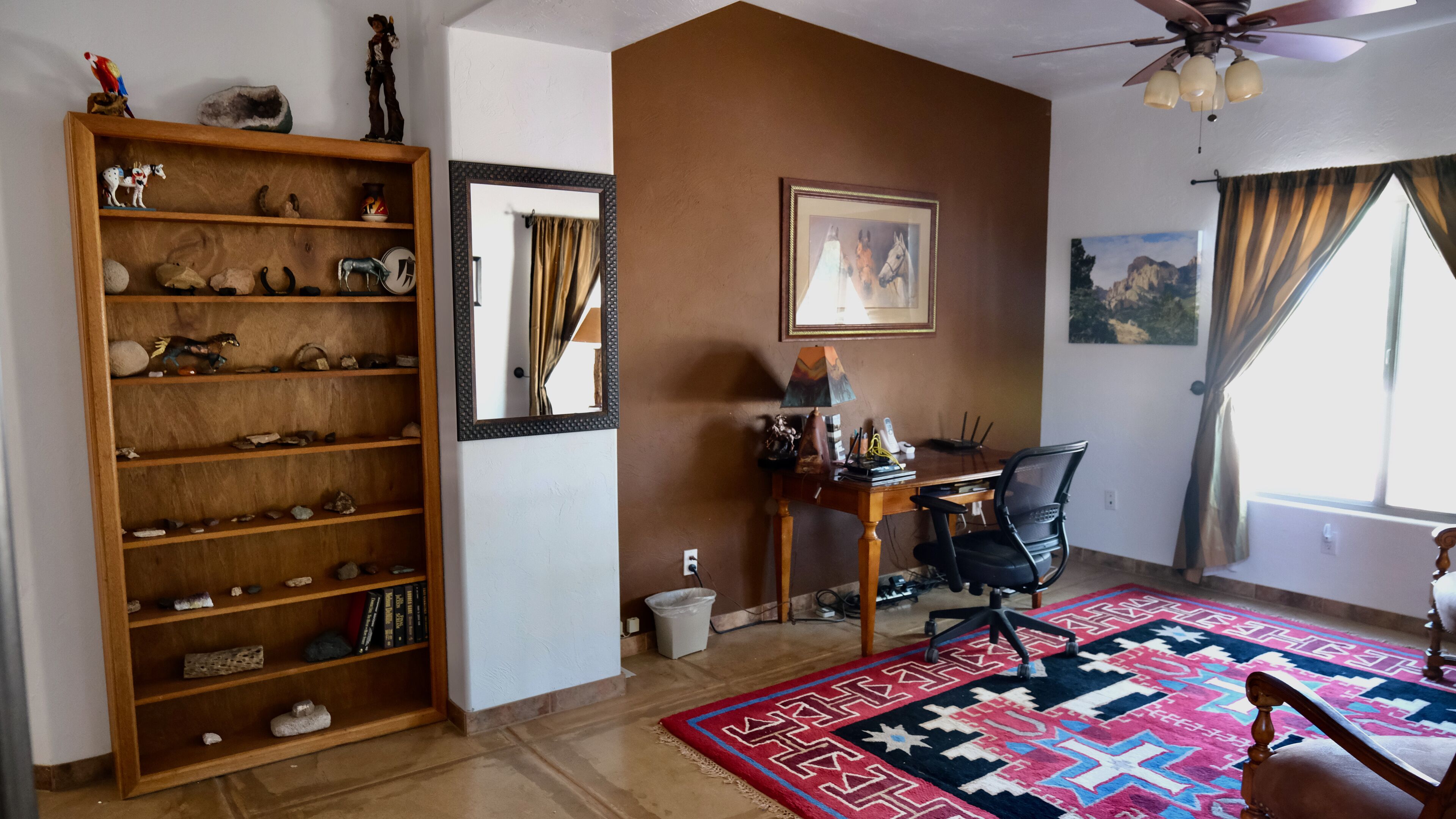 A room with a wooden bookshelf, a desk with a chair, a rug, a painting, and a ceiling fan.