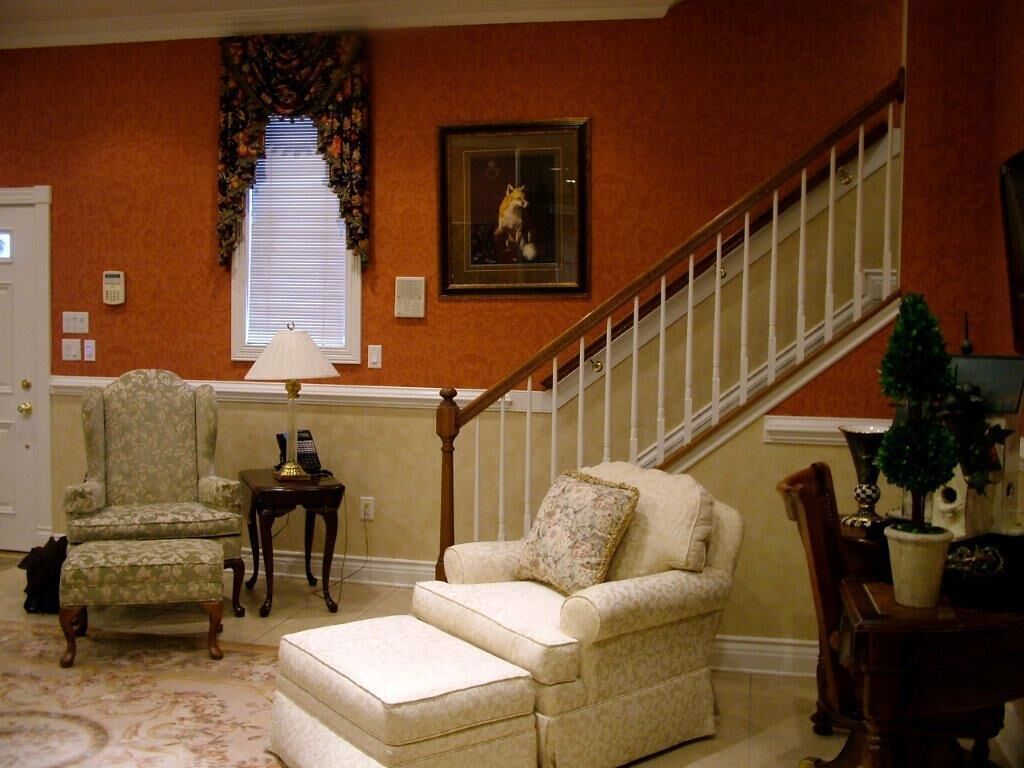 A living room with a staircase, a patterned armchair, a sofa, and a small table with a lamp.