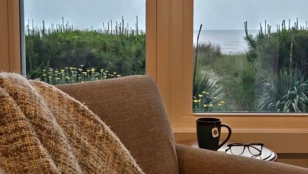 A room with a large window offering a view of a beach and dunes.