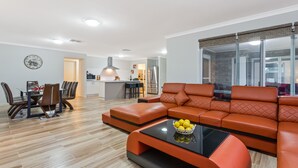 A modern living room with a sectional sofa, dining table, and chairs. There is a kitchen in the background with a refrigerator and a hanging light fixture. - Bulla Hill Villas- Melbourne Airport (Bulla)