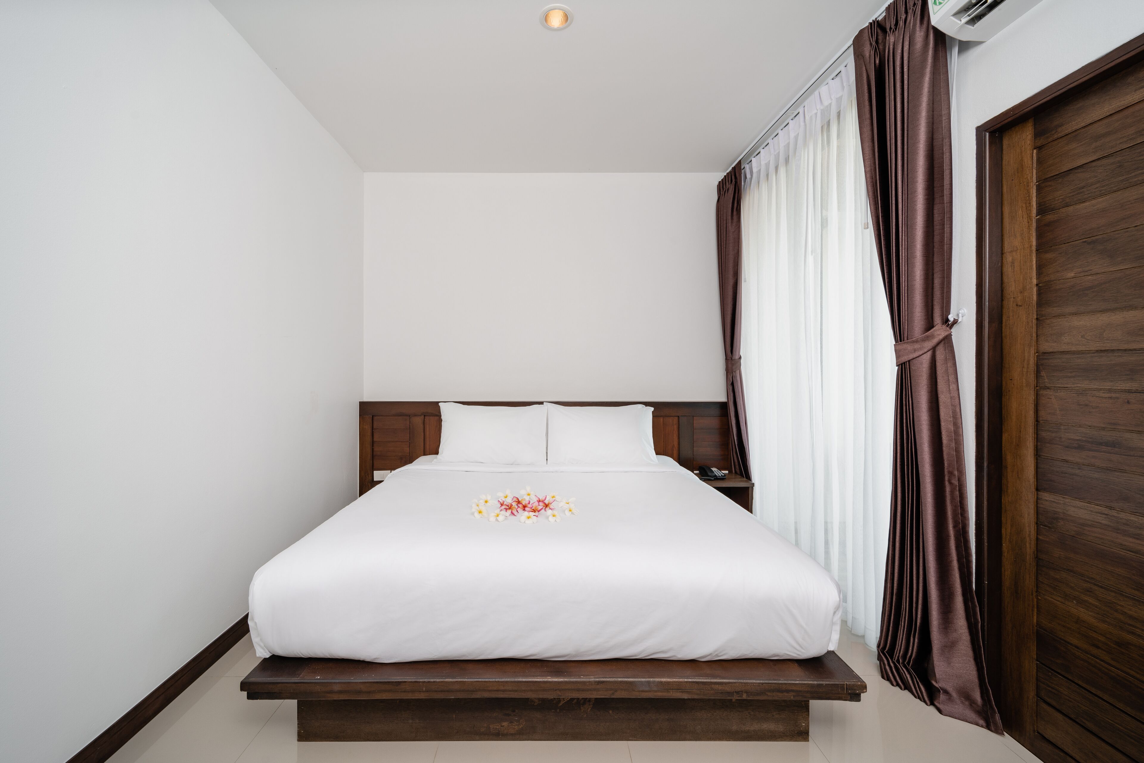 A neatly made bed with a white bedspread and a floral arrangement, a wooden headboard, a nightstand with a lamp, and a large wooden door.