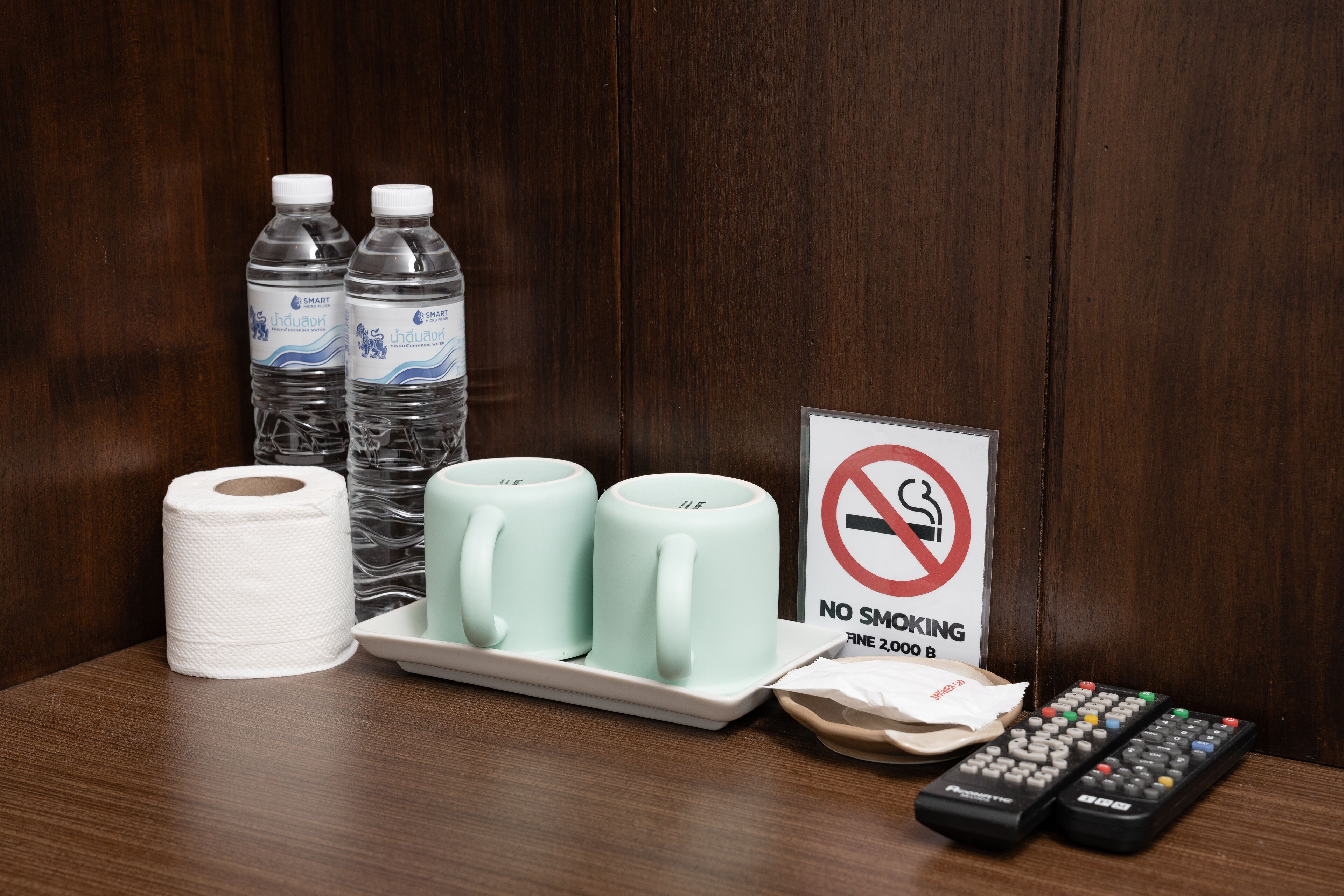 A hotel room with a no smoking sign, two water bottles, a roll of toilet paper, two light blue mugs, a tray with napkins, and two remote controls.