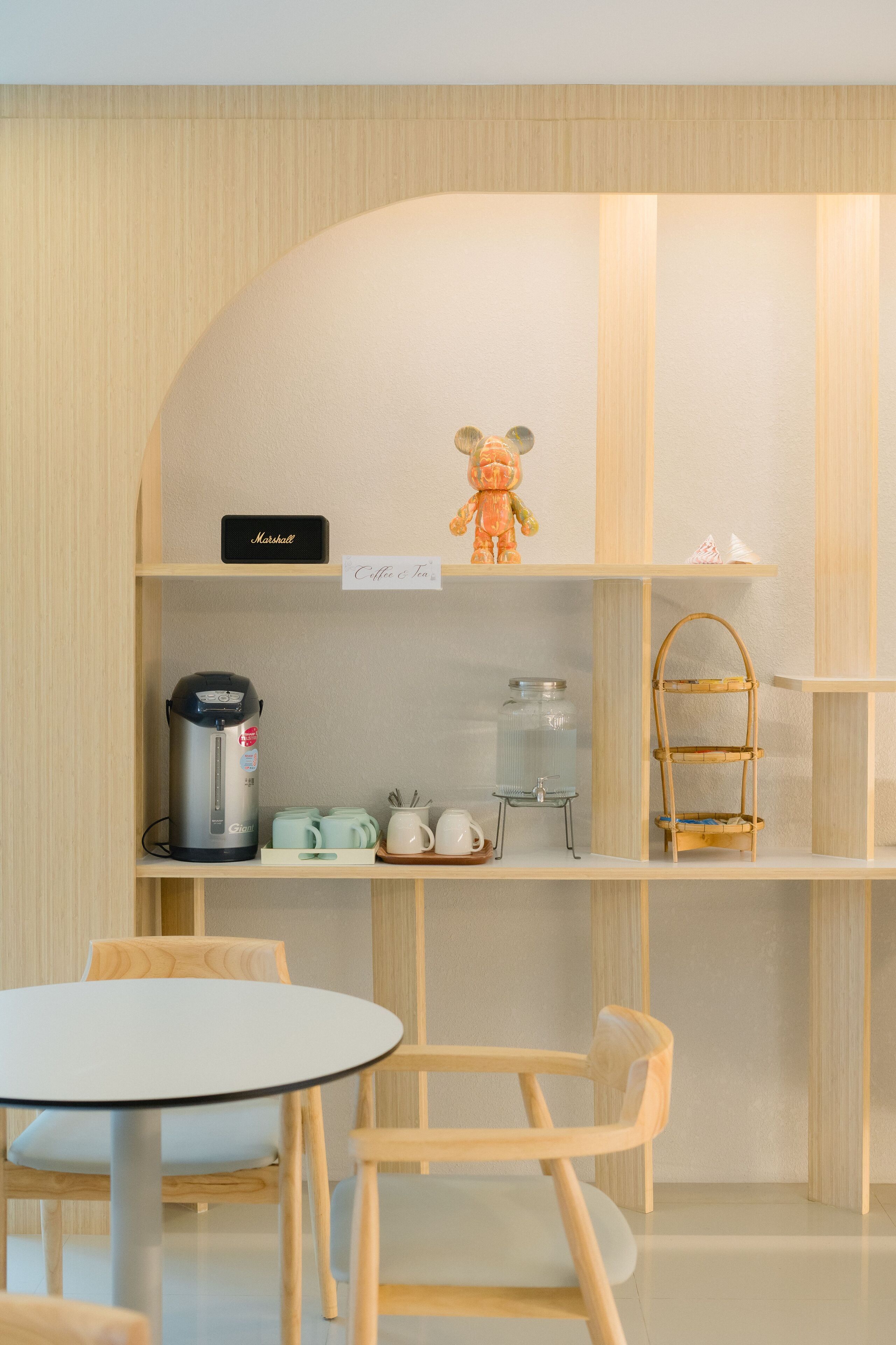 A compact, modern kitchenette with a coffee maker, tea set, and a small shelf with a teddy bear. There are two wooden chairs and a small table.