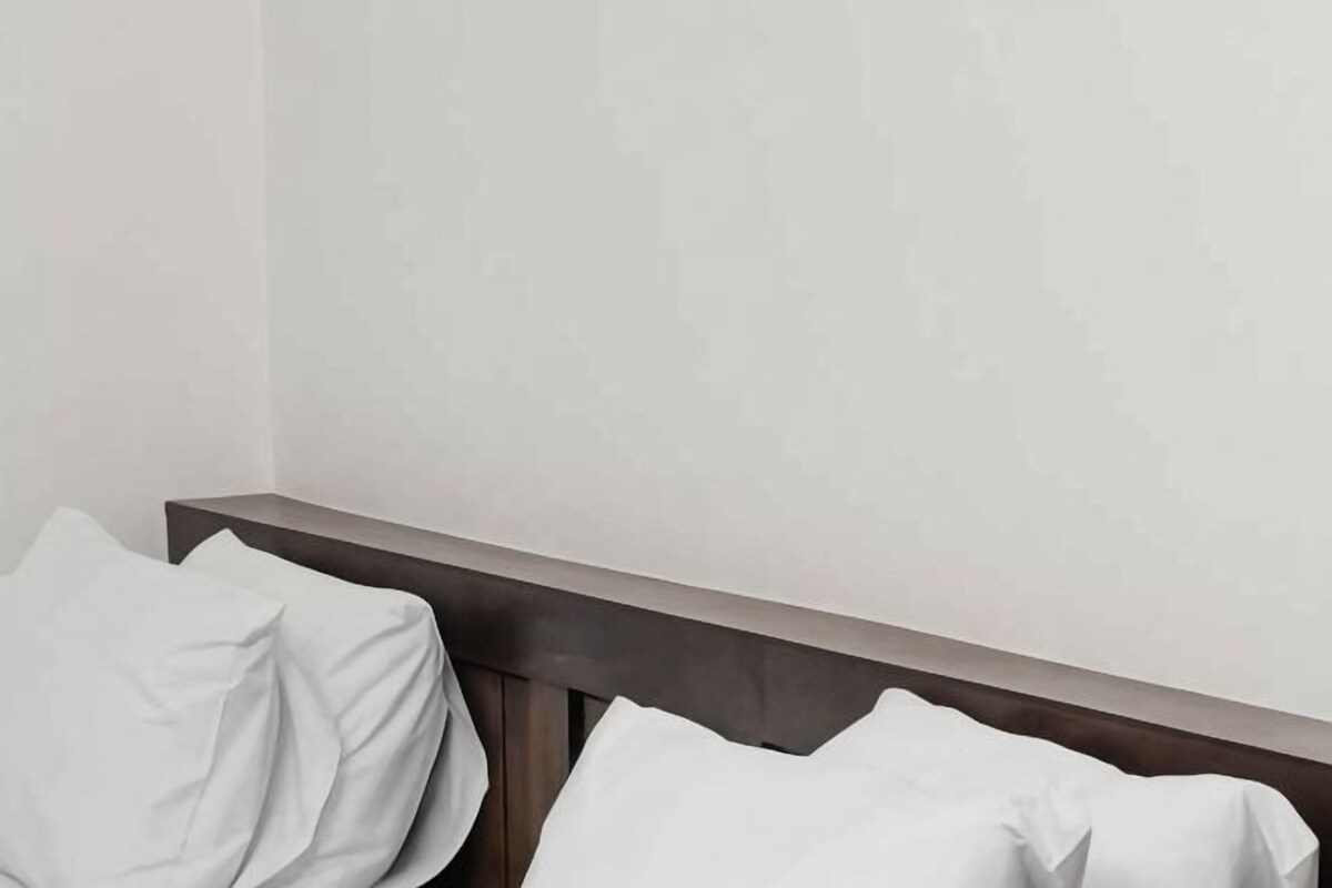 A room with a dark headboard against a plain wall, a white bedspread, and a white pillow.