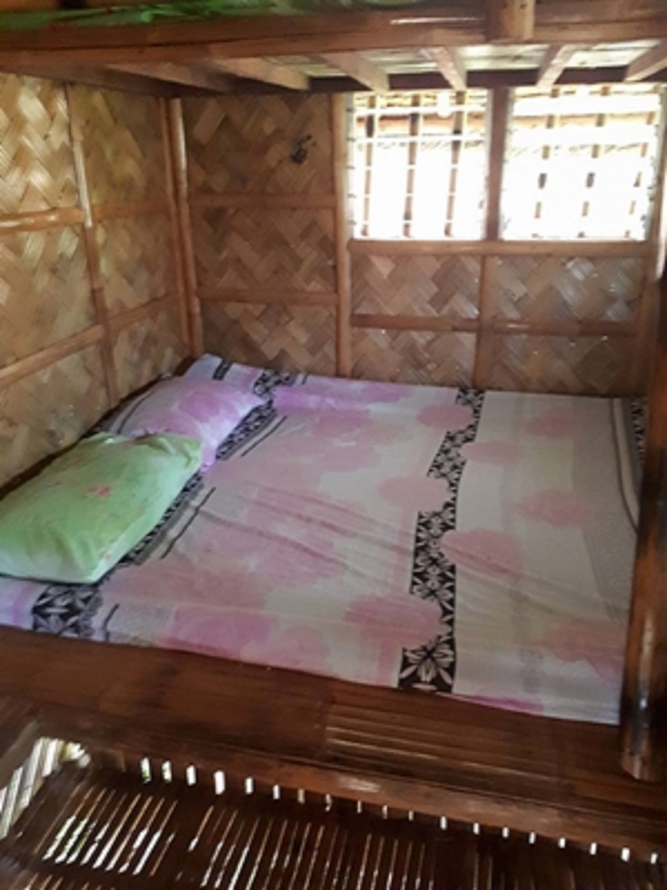 Una cama de madera con colcha estampada y almohada verde en una habitación con paredes de bambú y ventana.