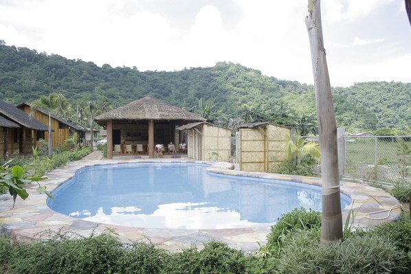 Una piscina con una estructura de techo de paja al fondo, rodeada de abundante vegetación y un paisaje montañoso.