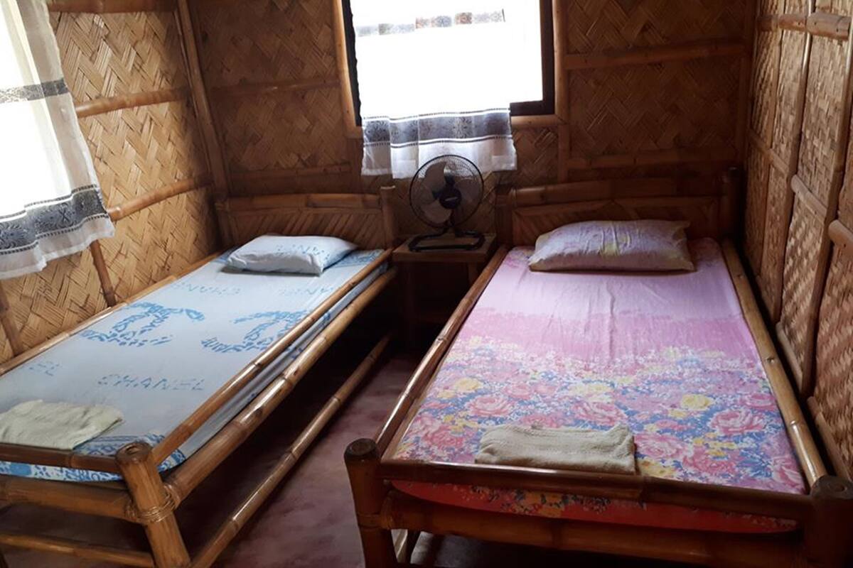 A room with two wooden bunk beds, a small bedside table with a fan, and a window with a curtain.